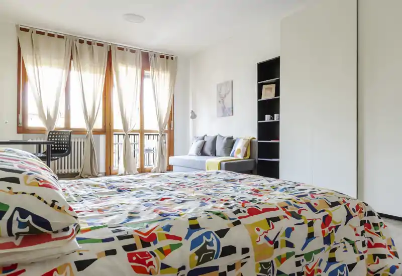 Camera da letto luminosa e moderna con copriletto colorato, grandi finestre con tende leggere, un piccolo divano e scaffalatura — perfetta per un alloggio allegro e ben illuminato.