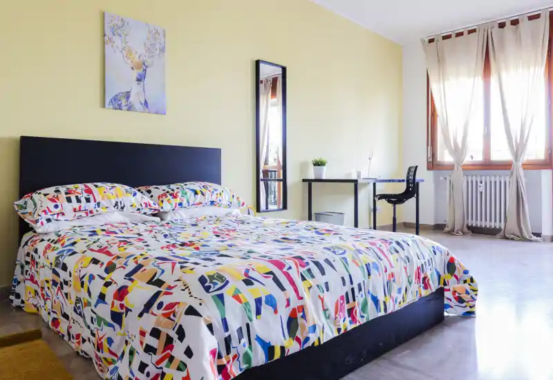 Camera da letto luminosa e di stile con un ampio letto matrimoniale con biancheria colorata, specchio a figura intera, scrivania e sedia vicino alla finestra soleggiata — ariosa e accogliente.