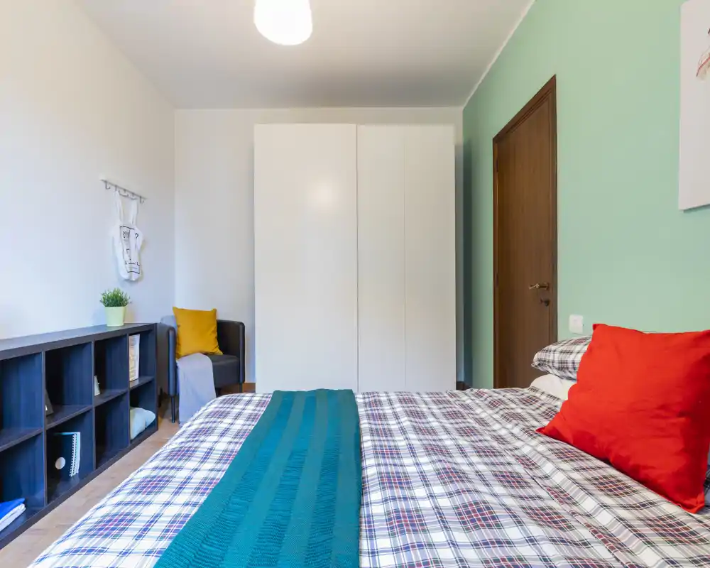 Camera da letto moderna e luminosa con copriletto a quadri, cuscini colorati e un ampio armadio bianco. La composizione mette in risalto un'area notte accogliente con spazio di stoccaggio ordinato e luce piacevole.