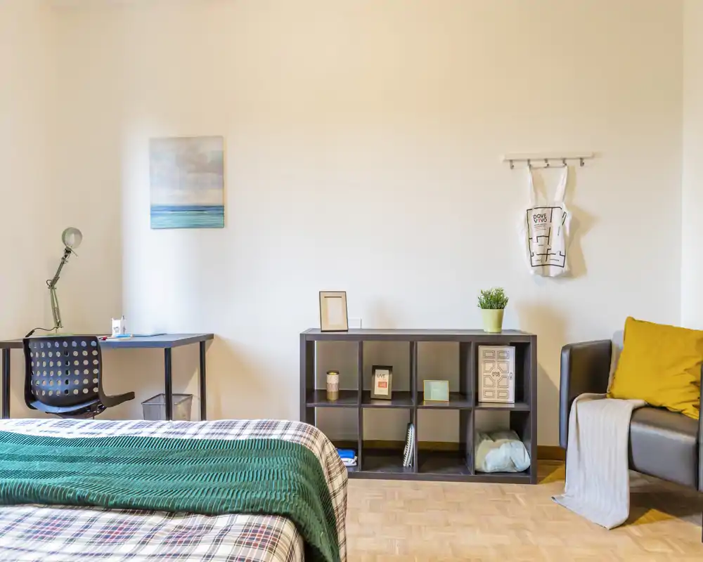 Camera da letto ben illuminata e preparata: letto con copriletto a fantasia e plaid verde, scrivania, libreria e poltrona accogliente — ambiente luminoso e invitante per annunci.