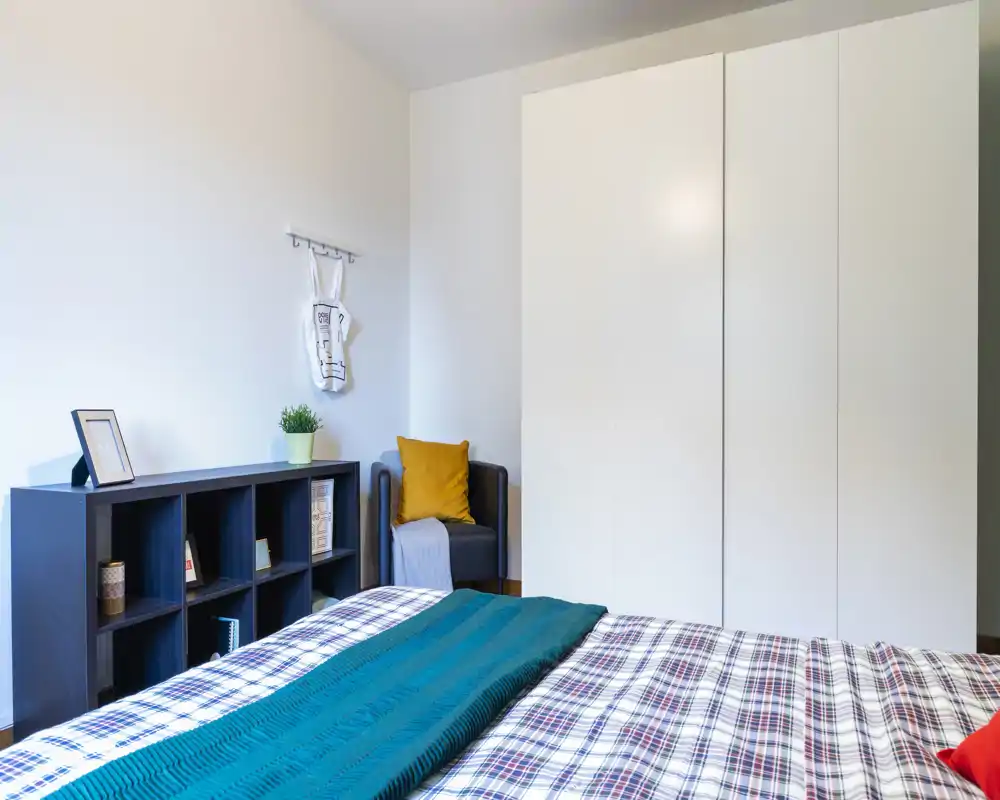 Camera da letto luminosa e ordinata con ampio letto in primo piano, armadio bianco a tutta altezza e una piccola poltrona con cuscino color senape. L'ambiente risulta fresco e accogliente grazie alle linee pulite, alle lenzuola a quadri e all'illuminazione naturale.