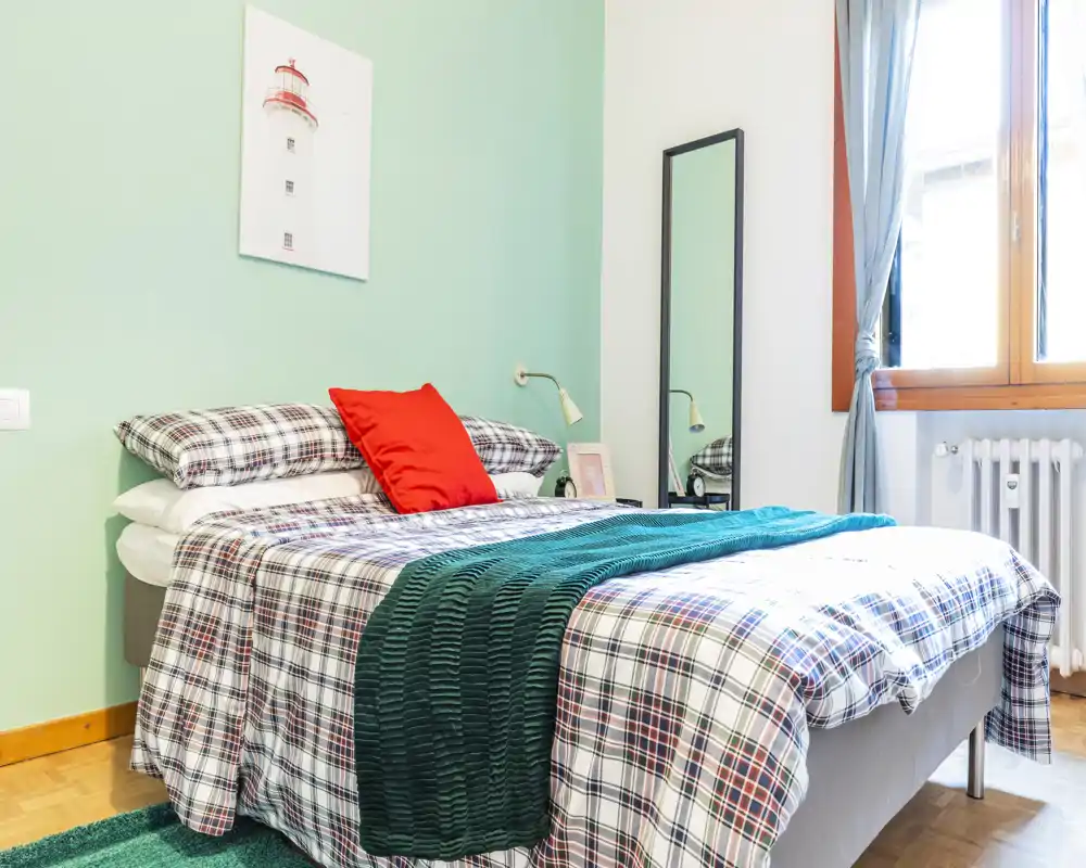 Camera da letto luminosa e curata con letto matrimoniale con lenzuola a quadri, plaid verde e cuscino rosso; luce naturale dalla finestra e arredi semplici creano un’atmosfera accogliente.