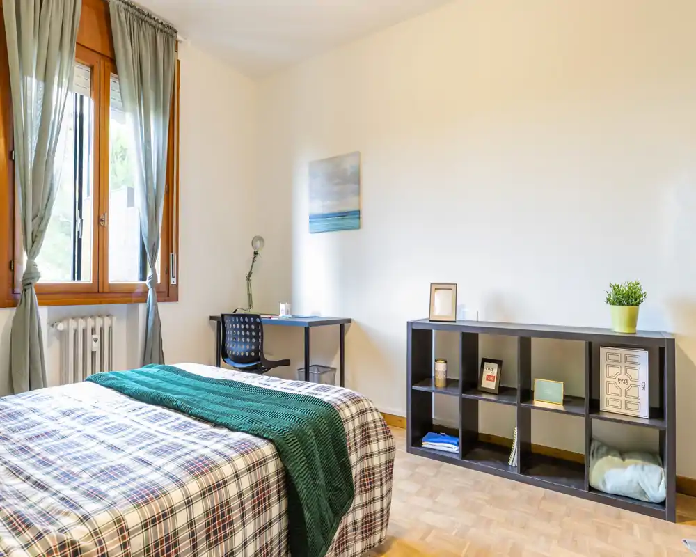 Camera da letto luminosa e ben arredata con un ampio letto con copriletto a fantasia e plaid verde, una scrivania vicino alla finestra e scaffalatura per riporre oggetti — perfetta per soggiorni confortevoli.