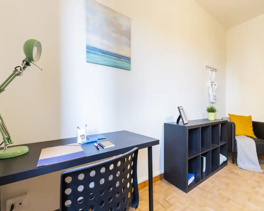 Area giorno luminosa e ordinata con una scrivania compatta, un mobile a cubi e un angolo divano accogliente con cuscino giallo e una lampada decorativa — perfetto per un piccolo appartamento.