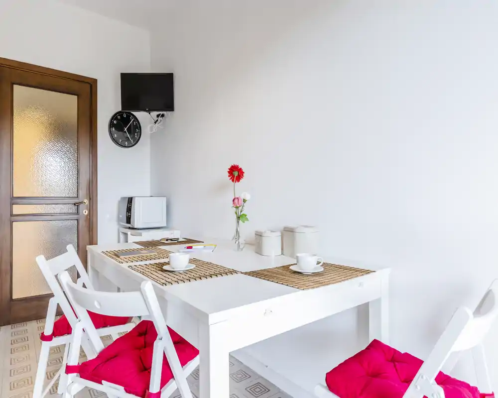 Zona pranzo luminosa e minimale con tavolo bianco, cuscini rossi e un semplice centrotavola floreale — perfetta per colazioni accoglienti o pasti leggeri.