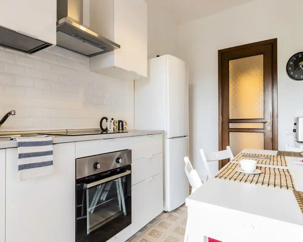 Cucina moderna e luminosa con mobili bianchi, forno incassato e frigorifero; un tavolo con tovagliette crea un angolo colazione accogliente.