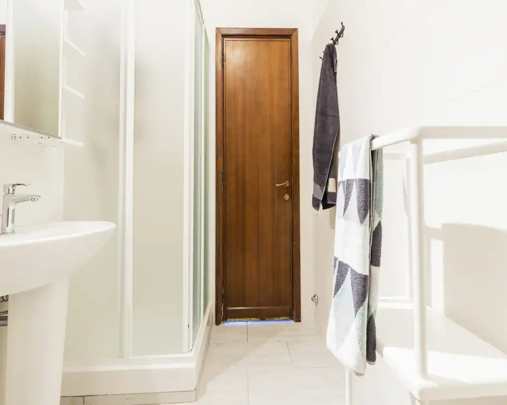 Bagno luminoso e moderno con cabina doccia angolare, lavabo a colonna, portasciugamani e porta in legno; piastrelle bianche e luce naturale morbida.
