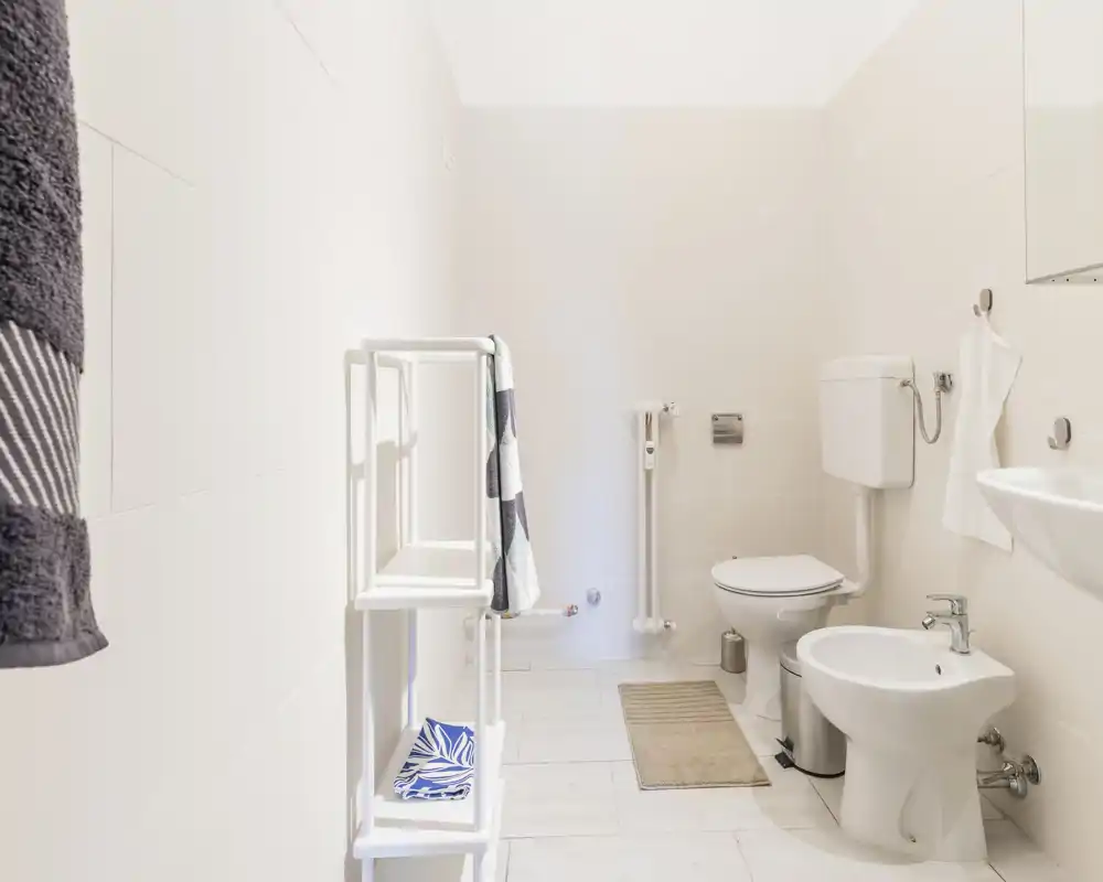 Bagno moderno e luminoso con wc, bidet e lavabo a parete; piastrelle chiare e scaffalatura semplice per un aspetto pulito e minimale.