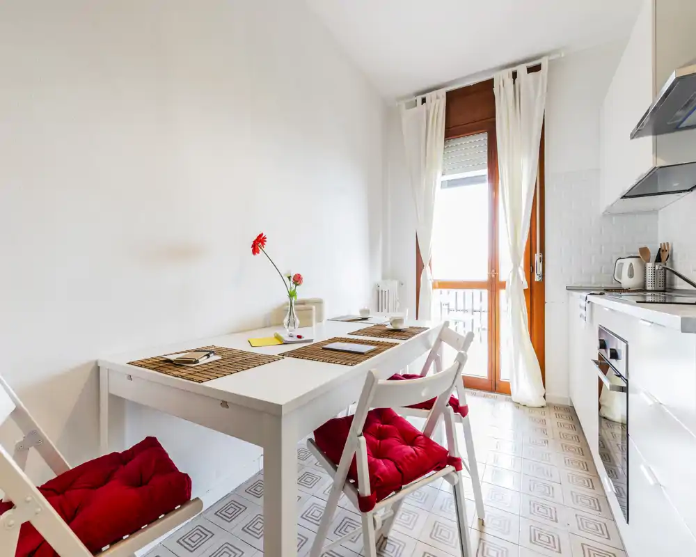 Cucina compatta e luminosa con tavolo bianco, sedie con cuscini rossi e porta sul balcone che fa entrare luce naturale — ideale per un piccolo appartamento.