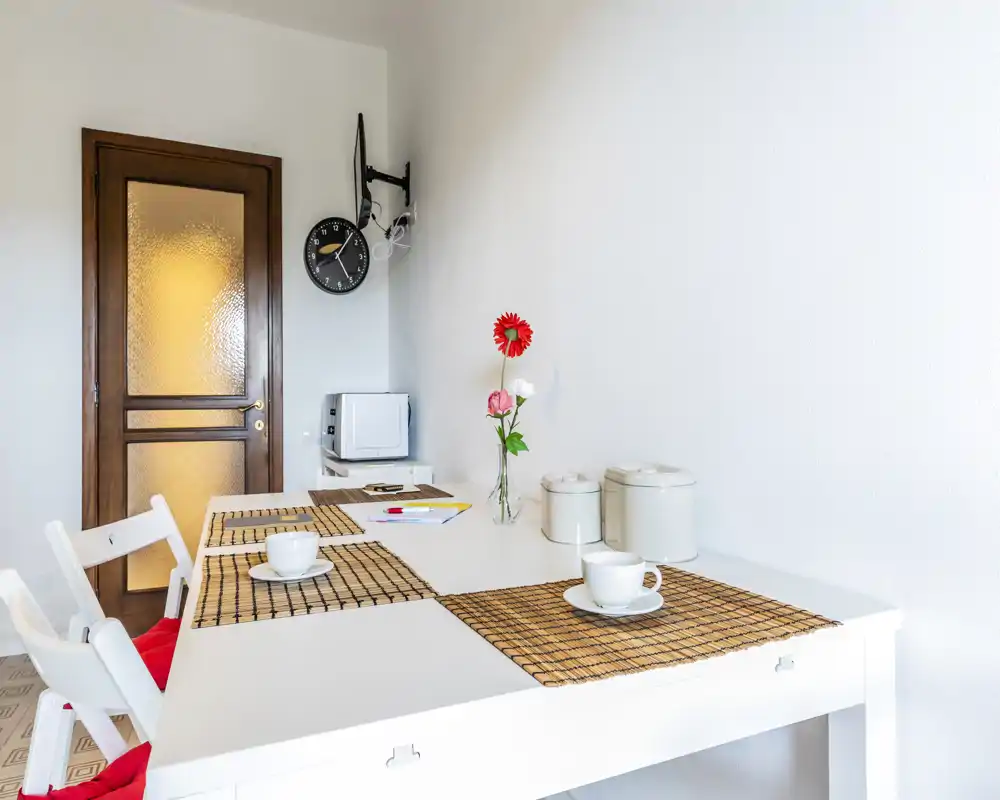 Zona pranzo luminosa e minimal con tavolo bianco, tovagliette, tazze e un piccolo vaso di fiori. Lo spazio è ordinato e accogliente, ideale per pasti informali.