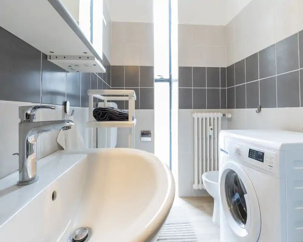 Bagno luminoso e moderno con lavabo, rubinetto, termosifone e lavatrice a carica frontale; piastrelle neutre e luce naturale da una finestra alta.
