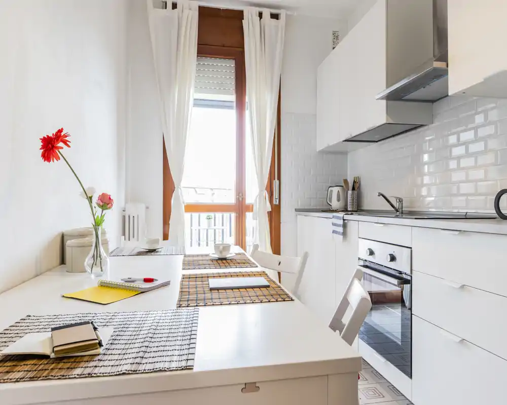 Cucina moderna e luminosa con tavolo da pranzo/lavoro, mobili bianchi, forno integrato e dettagli decorativi come vaso di fiori e tovagliette, perfetta per un appartamento compatto.
