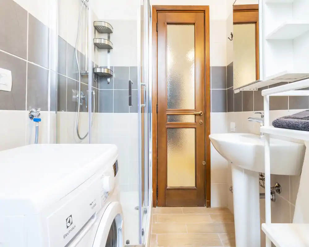 Bagno luminoso e moderno con box doccia in vetro, lavabo a colonna, lavatrice e porta in legno—spazio pulito e ben illuminato ideale per gli annunci.