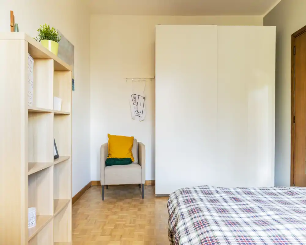 Dormitorio luminoso y de estilo sencillo con cama de cuadros, un sillón compacto con cojines coloridos y un gran armario blanco — aireado, ordenado y perfecto para un anuncio de alquiler.