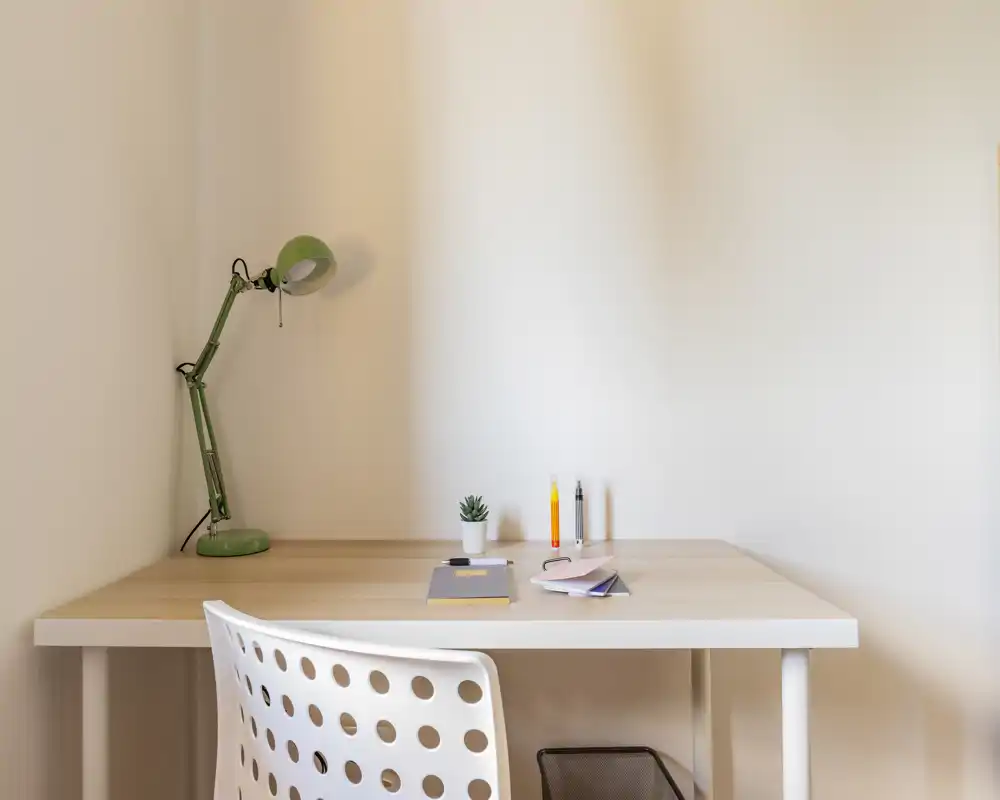 Rincón de escritorio ordenado con mesa de madera minimalista, silla blanca perforada, lámpara verde ajustable y útiles: espacio de trabajo luminoso y tranquilo, perfecto para estudiar o trabajar desde casa.