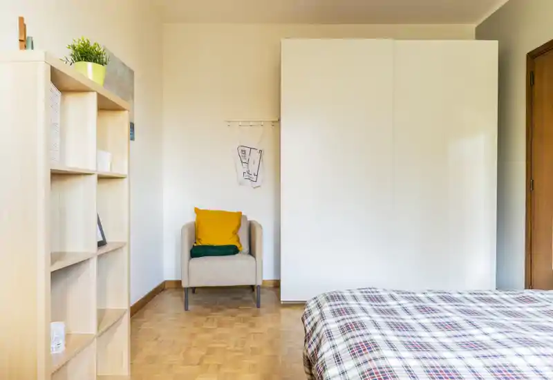 Dormitorio luminoso y de estilo sencillo con cama de cuadros, un sillón compacto con cojines coloridos y un gran armario blanco — aireado, ordenado y perfecto para un anuncio de alquiler.