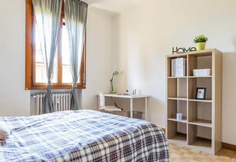 Dormitorio luminoso y ordenado con cama a cuadros, escritorio junto a la ventana y estantería abierta — ideal para estancias cortas o largas.