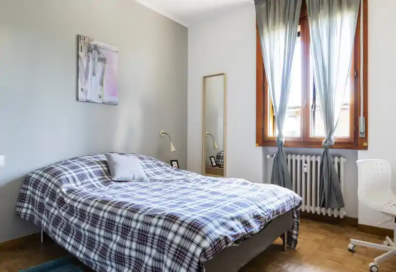 Dormitorio luminoso y ordenado con una cama doble con edredón de cuadros, una lámpara de noche y una gran ventana con cortinas que aporta abundante luz natural.