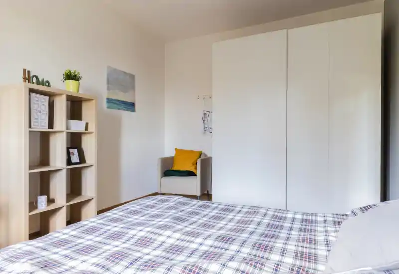 Dormitorio luminoso y ordenado con colcha de cuadros en primer plano, armario blanco y un sillón con cojín colorido — una zona de descanso tranquila y bien presentada.
