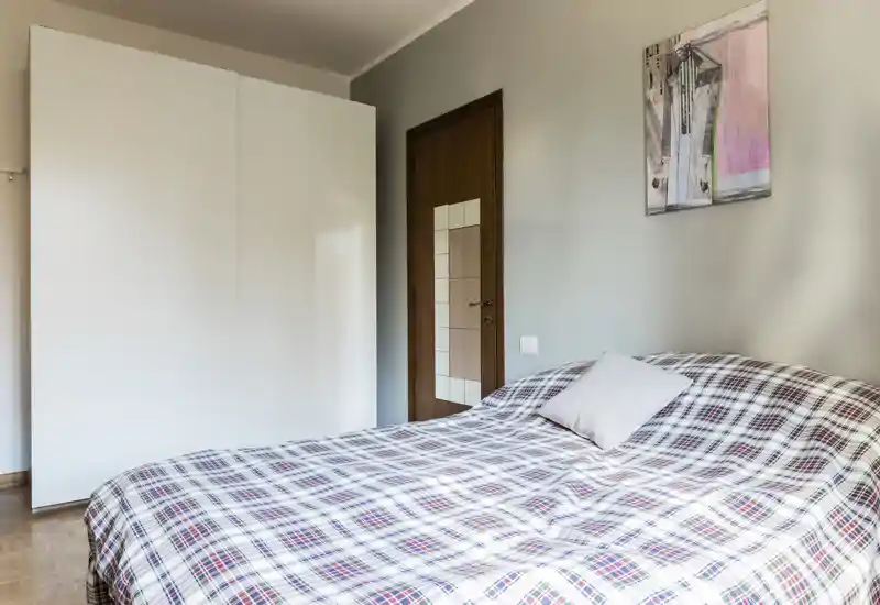 Dormitorio luminoso y ordenado con una gran cama con colcha de cuadros, un armario blanco y una puerta de madera; estilo sencillo y contemporáneo apto para anuncios.