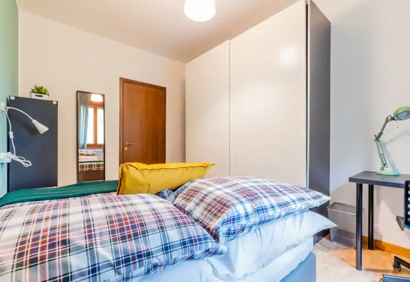 Bright, well-styled bedroom with a large bed dressed in plaid linens, a mustard cushion and a wardrobe along the wall. The room shows a bedside desk and lamp, wooden door and a standing mirror reflecting additional natural light.