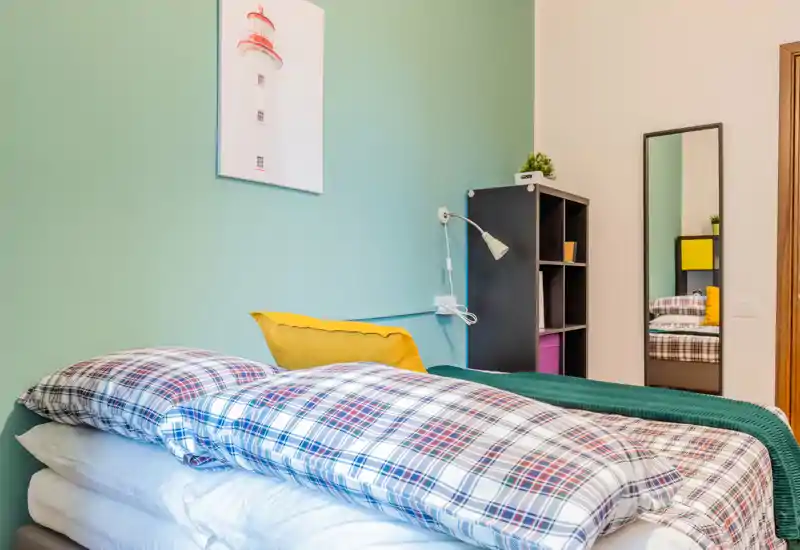 Bright, cozy bedroom with a patterned duvet, green accent wall and a full single/double bed in the foreground; shelving and a full-length mirror add practical storage and depth.