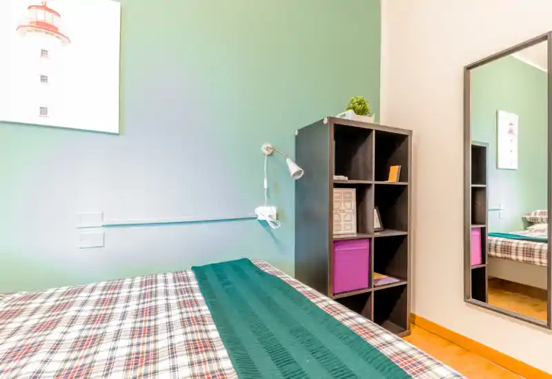 Chambre lumineuse et soignée avec couette à carreaux, jeté vert et étagère cubique ; un miroir pleine longueur apporte de la profondeur.