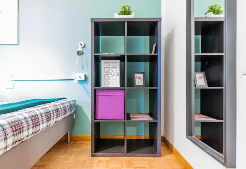 Angolo camera da letto compatto e ben illuminato con un letto singolo e una libreria a cubi; arredo moderno e semplice con piumone a quadri e uno specchio alto che amplia visivamente lo spazio.