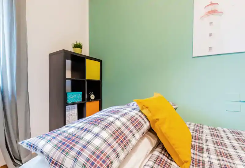 Bright, modern bedroom corner with plaid bedding and a vibrant yellow cushion; simple shelving and soft green accent wall create a fresh, welcoming look.