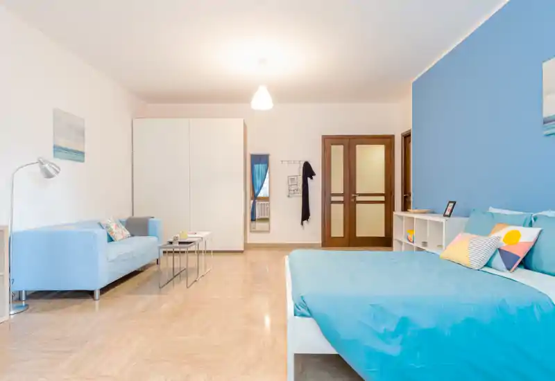 Camera da letto moderna e luminosa con tonalità azzurre. Il letto è completamente visibile con cuscini coordinati e un mobiletto a giorno; un piccolo divano e l'armadio completano la disposizione accogliente.