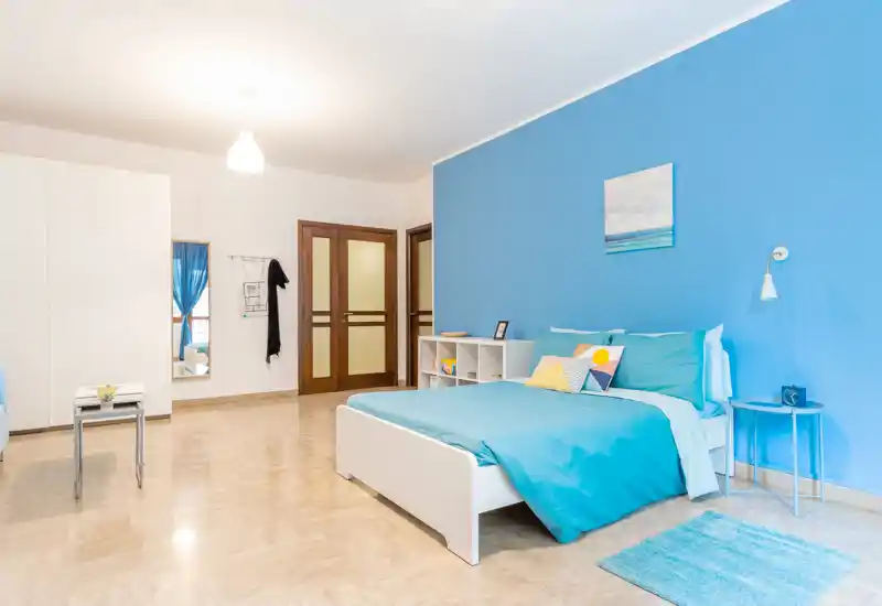 Camera da letto luminosa e moderna con parete accent in turchese e letto matrimoniale ben rifatto. Tessuti morbidi, cuscini colorati e un piccolo comodino rendono l’ambiente accogliente e curato.