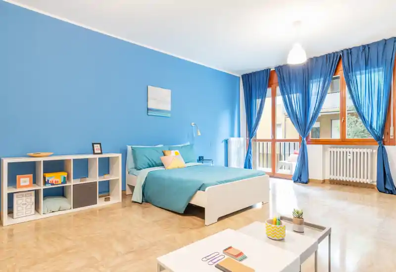 Camera da letto luminosa e moderna con letto matrimoniale con lenzuola turchesi, ampie finestre con tende blu e pavimento chiaro che crea un’atmosfera ariosa e contemporanea.