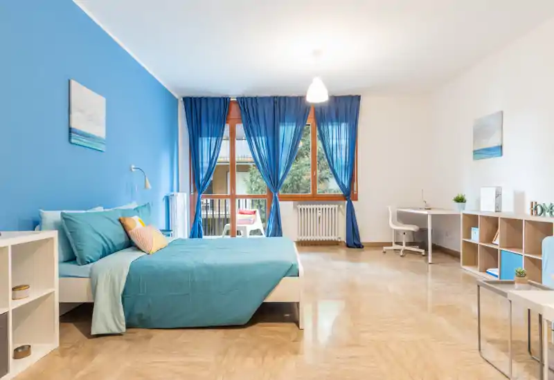 Camera da letto luminosa e moderna con parete accent color turchese, ampia finestra con tende blu e letto matrimoniale ben rifatto. Presente una scrivania e scaffalature aperte, perfetta per soggiorni comodi.