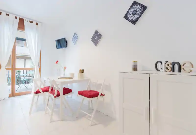 Angolo pranzo luminoso e minimal con un piccolo tavolo bianco, quattro sedie pieghevoli con cuscini rossi e una porta sul balcone che illumina l'ambiente con luce naturale.
