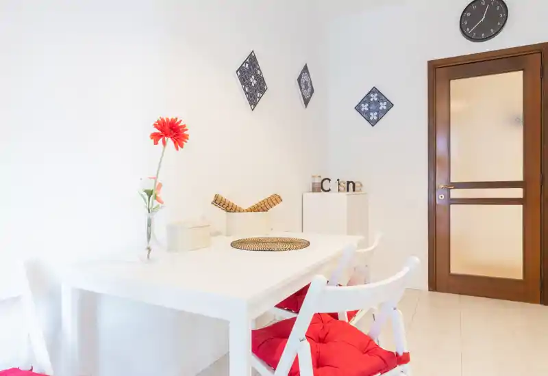 Zona pranzo luminosa e minimal con tavolo bianco, sedie con cuscini rossi e decorazioni semplici, ideale per un monolocale o un piccolo appartamento.