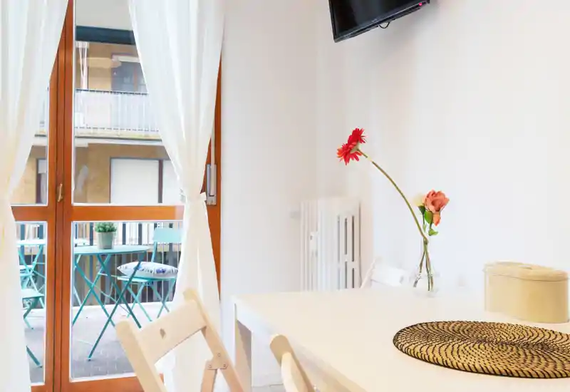 Zona giorno luminosa e minimal con tavolo bianco, sedie semplici, fiori decorativi e porta finestra che dà su un piccolo balcone. Luce naturale e tessuti morbidi creano un ambiente accogliente.