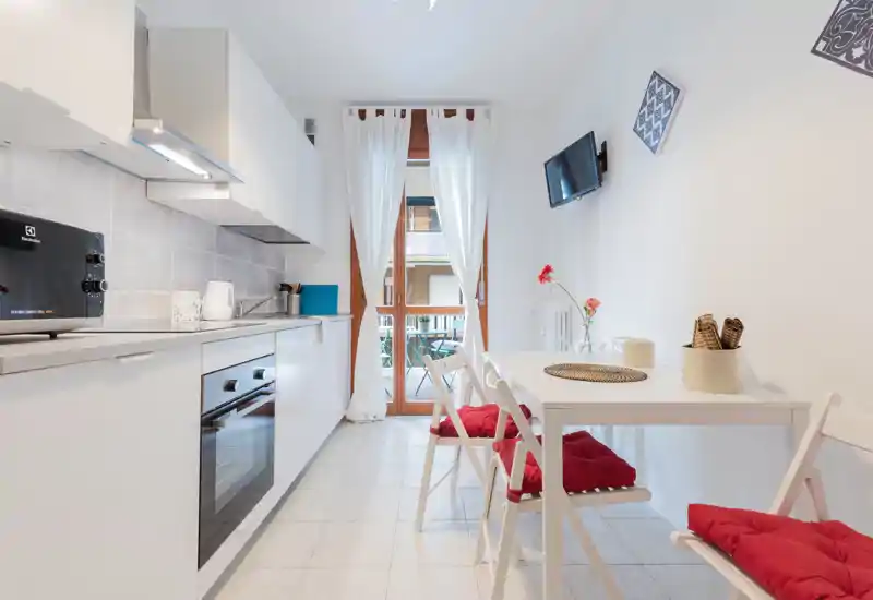 Cucina angolo cottura luminosa e moderna con mobili bianchi, forno incassato, piccoli elettrodomestici e un tavolo da pranzo con cuscini rossi; porta balcone che illumina naturalmente.