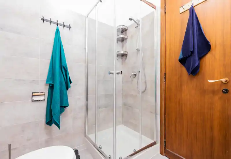 Bagno moderno e luminoso con doccia a vetri, piastrelle chiare e porta in legno; asciugamani appesi su ganci e disposizione compatta, ideale per un annuncio di affitto.