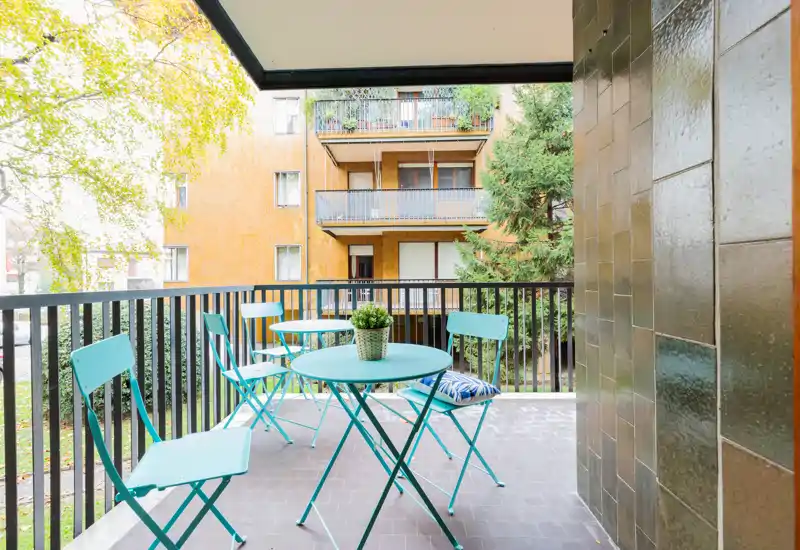 Balcone soleggiato arredato con tavolino rotondo, quattro sedie pieghevoli turchesi abbinate e una pianta decorativa — perfetto per il caffè mattutino o per rilassarsi all'aperto.