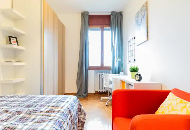 Bright, modern bedroom with a patterned bedspread, a desk by the window and a vivid red armchair adding a pop of color — ideal for students or short-term rentals.