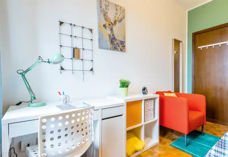Bright, modern bedroom corner with a white study desk, mint green lamp and a vibrant orange armchair. The tidy shelving and decorative accents create a stylish, cozy spot ideal for studying or relaxing.