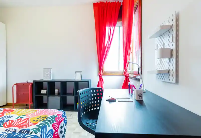 Bright, colorful bedroom corner with a patterned bedspread, workspace with desk and chair, shelving unit and a window with red curtains letting in natural light.