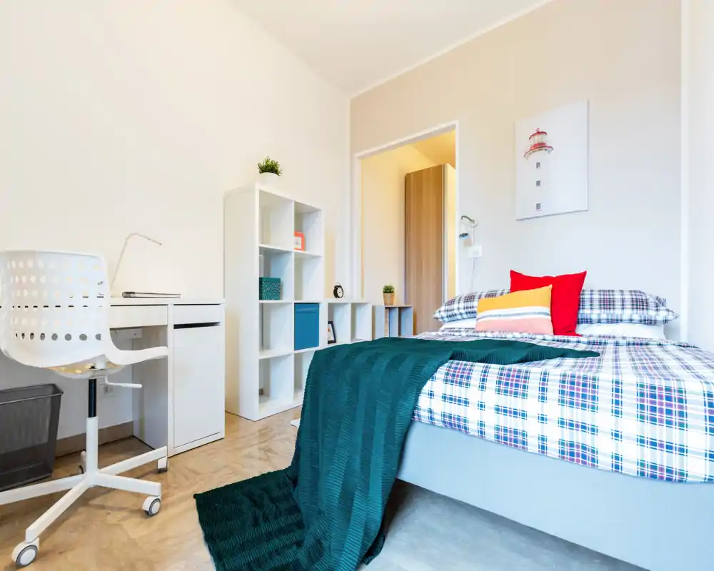 Camera da letto luminosa e ben arredata con letto matrimoniale con lenzuola a quadri, cuscini colorati e plaid; presente una scrivania e scaffalatura ideale per studio o deposito.