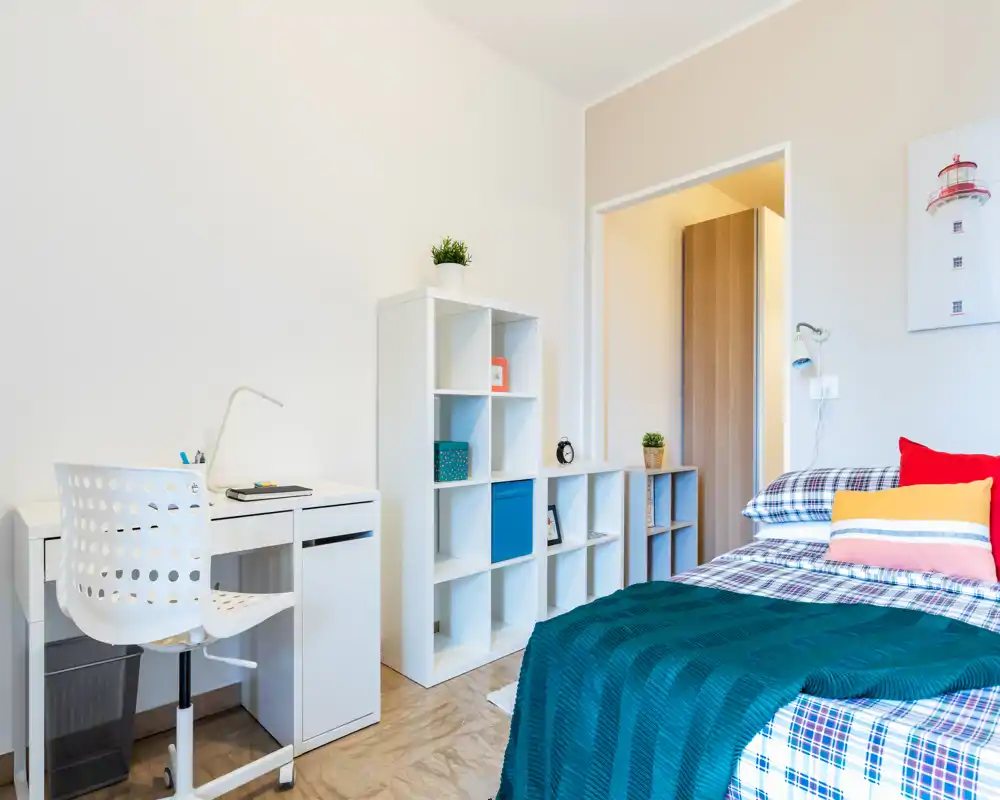 Camera da letto luminosa e moderna con letto ben rifatto, cuscini colorati e una coperta verde acqua; presente una scrivania bianca e scaffalatura modulare perfetta per studio o ripostiglio.