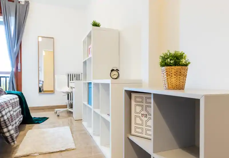 Camera da letto moderna e luminosa con letto ordinato, scaffalature aperte, un tappeto accogliente e una scrivania vicino alla finestra — perfetta per annunci di affitti brevi.
