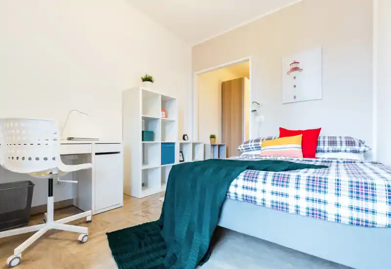 Camera da letto luminosa e ben arredata con letto matrimoniale con lenzuola a quadri, cuscini colorati e plaid; presente una scrivania e scaffalatura ideale per studio o deposito.