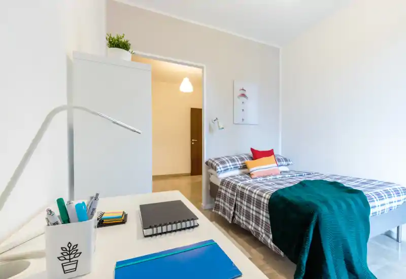 Camera da letto singola luminosa e ordinata con copriletto a quadri, cuscini colorati e un plaid verde. Una scrivania compatta in primo piano crea un angolo studio pratico; pareti neutre e arredi semplici donano un aspetto moderno e rilassante.