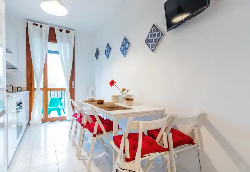 Bright, compact kitchen-dining area with a white table, folding chairs with red cushions, and a balcony door letting in natural light — ideal for casual meals.