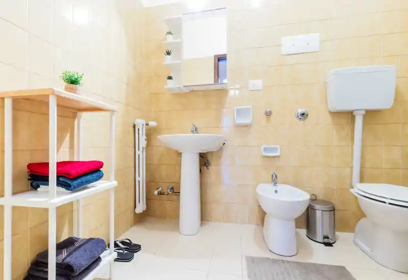 Bright, well-maintained bathroom with pedestal sink, bidet and toilet; neutral beige tiles and tidy shelving with folded towels.