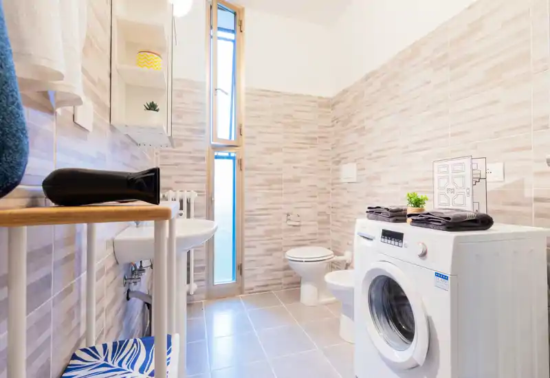 Bright, modern bathroom with tiled walls, a pedestal sink, toilet and a front-loading washing machine; light, neutral tones and a tall narrow window provide good natural light.